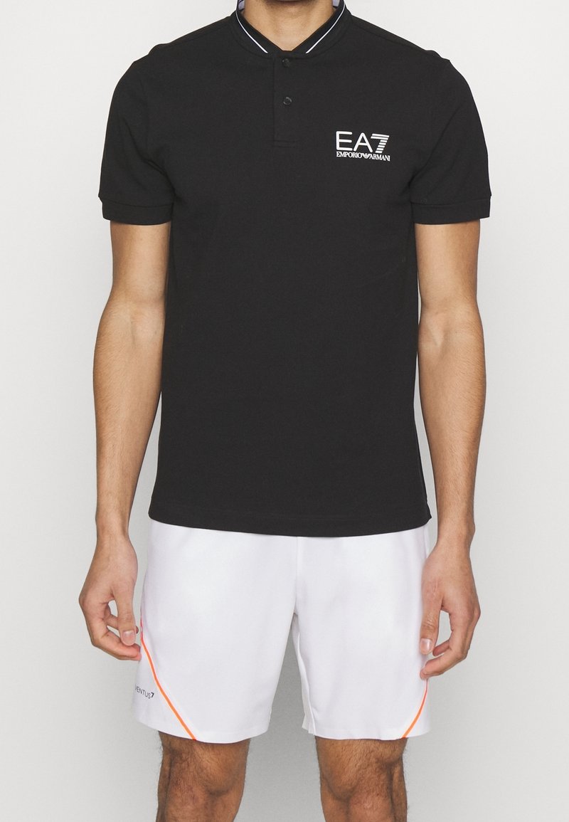 Homme portant un polo noir EA7 Emporio Armani et un short blanc avec des détails orange, debout devant un fond uni.