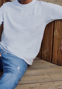 Světle šedá mikina s malými modrými logy Levi's, kulatým výstřihem a volným střihem, kombinovaná s modrými zničenými džíny na dřevěném pozadí.