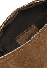 Bolso de ante marrón con cremallera plateada parcialmente abierta, mostrando el interior de tela negra y una etiqueta de cuero marrón con letras en relieve.
