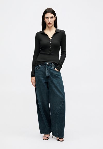 Femme aux longs cheveux foncés portant un haut noir à manches longues boutonné, un jean bleu foncé à jambes larges et des sandales à talons hauts noires.