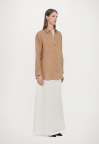 Light brown linen shirt with a collar, button front, and chest pocket, paired with a long, white satin skirt and black shoes.