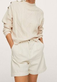 Pull beige clair en maille torsadée avec col côtelé et boutons aux épaules, associé à un short plissé de couleur crème.