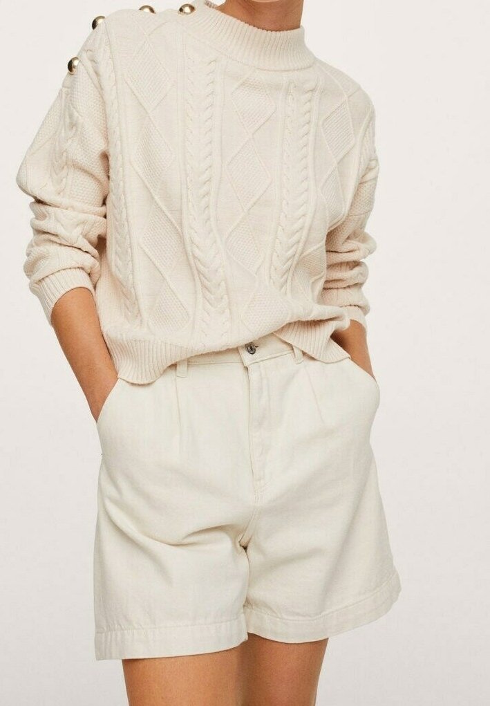 Pull beige clair en maille torsadée avec col côtelé et boutons aux épaules, associé à un short plissé de couleur crème.