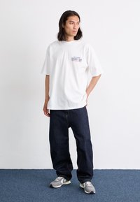 White cotton t-shirt with a purple and blue graphic, paired with dark blue denim pants. Model wears grey sneakers with black accents.