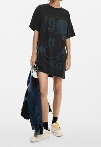 Czarna, oversize sukienka t-shirt z niebieską grafiką przedstawiającą postać z kreskówki oraz tekstem. Zestawiona z białymi sneakersami i dżinsową kurtką.