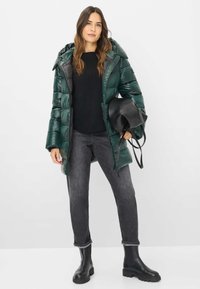 Grøn pufferjakke med en skinnende overflade, forsynet med hætte. Båret over en sort top og parret med grå jeans og sorte ankelstøvler.
