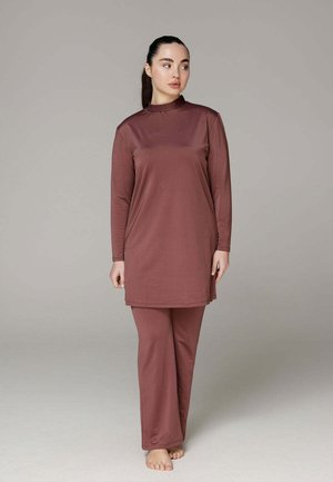 Femme debout pieds nus portant une tunique bordeaux à manches longues et un pantalon large assorti devant un fond gris uni.