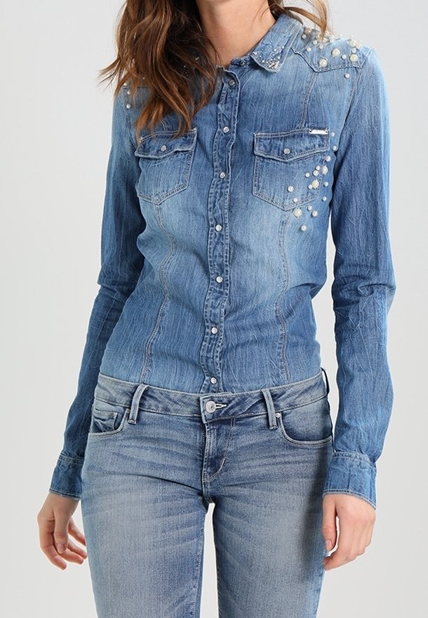 Femme portant une chemise en denim bleu ajustée avec des ornements en perles sur l'épaule et la poitrine, assortie à un jean bleu.