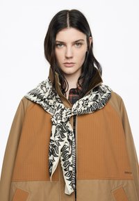 Una mujer con cabello oscuro lleva una chaqueta marrón con paneles naranjas y una bufanda de estampado animal en blanco y negro atada alrededor del cuello.