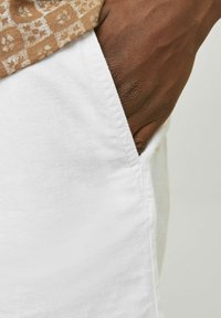 Shorts en lin blancs avec une poche latérale, dotés d'une texture douce. Une main brune est placée près de la poche, partiellement obscurcie.