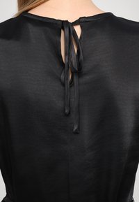 Czarna bluzka z wycięciem w kształcie klucza z tyłu, zabezpieczona wiązanymi satynowymi wstążkami. Materiał ma gładką, lekką teksturę.