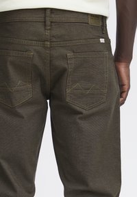 Jeans de mezclilla marrones con dos bolsillos traseros que presentan detalles de costura en forma de montaña y una pequeña etiqueta de la marca en la cinturilla.