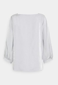 Blouse blanche à manches longues avec un col bateau et des manches légèrement bouffantes, vue de dos sur un fond uni.