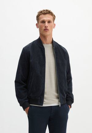 Junger Mann mit lockigem blondem Haar, der eine dunkelblaue Bomberjacke über einem hellbeigen Pullover trägt und mit den Händen in den Taschen vor weißem Hintergrund steht.