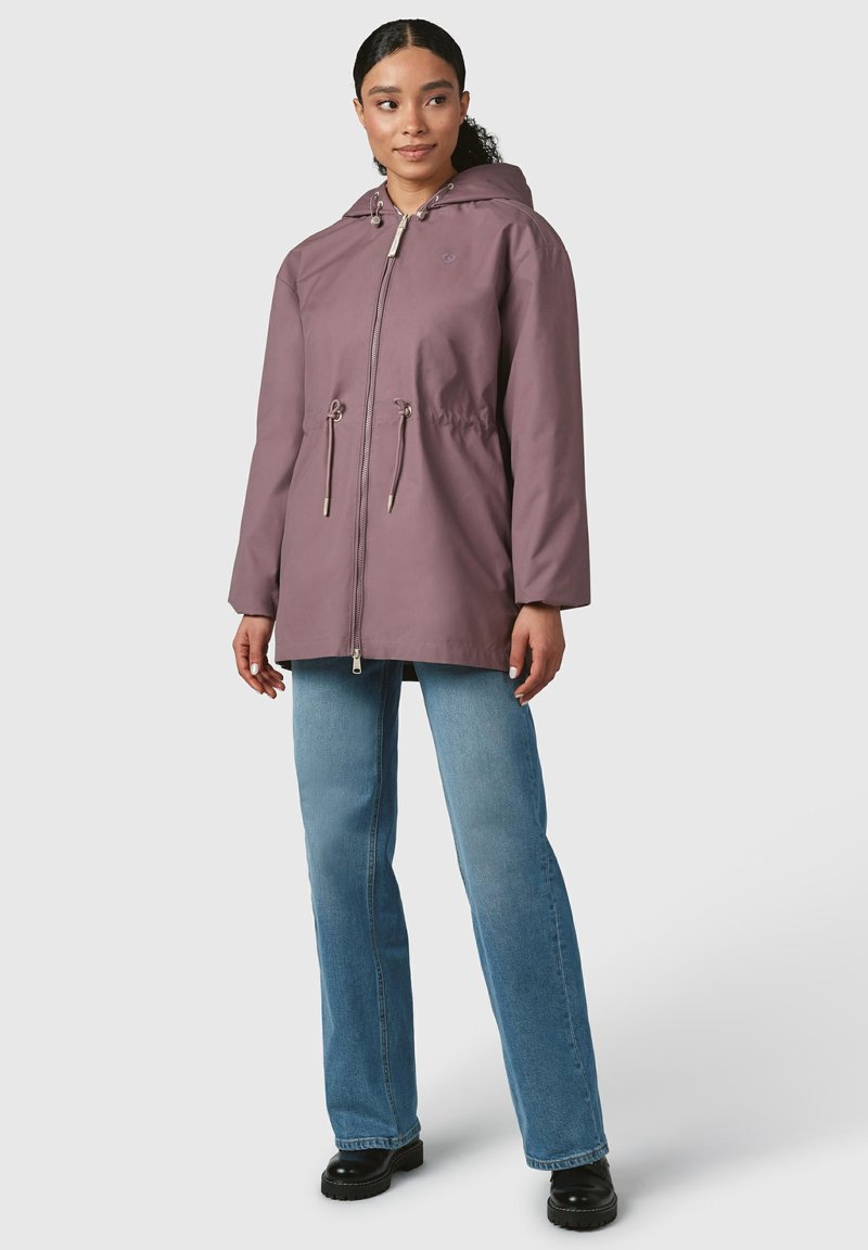 Giacca impermeabile di colore viola chiaro con cappuccio, stringhe regolabili e zip frontale. Indossata sopra jeans blu e stivali platform neri.