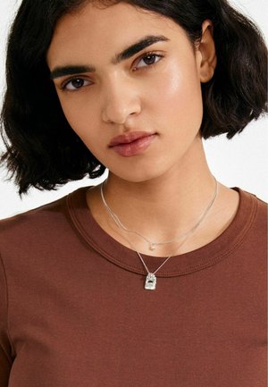 Silberne, geschichtete Halsketten mit einem strukturierten Anhänger und einer kleinen Perle, getragen über einem braunen, eng anliegenden Shirt mit rundem Ausschnitt.