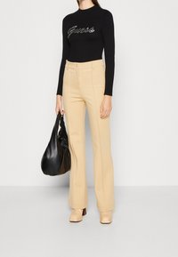 Schwarzes Langarmoberteil mit silbernem "Guess"-Logo, kombiniert mit hellbeigen, hochgeschnittenen Hosen. Das Model hält eine schwarze, lässige Handtasche.