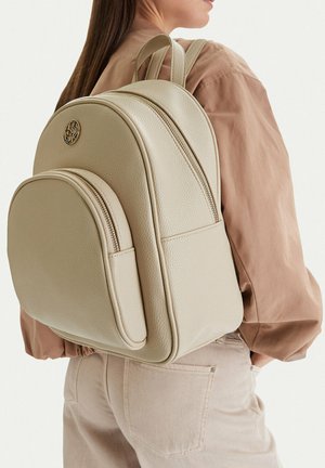Mujer con mochila de cuero beige con bolsillo delantero y logo dorado, combinada con camisa marrón claro y pantalones crema, vista desde atrás.
