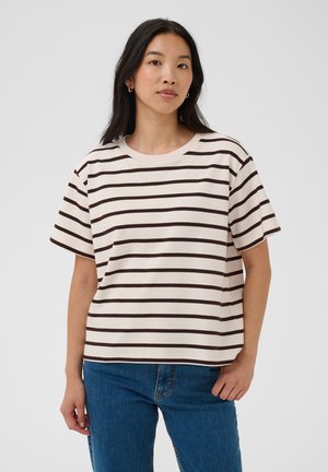 Jeune femme aux cheveux foncés portant un t-shirt blanc à manches courtes avec des rayures horizontales noires et un jean bleu, debout devant un fond uni.
