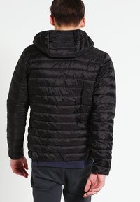 Mann in schwarzer gesteppter Kapuzenjacke und dunkler Hose, von hinten gesehen, vor einem weißen Hintergrund stehend.