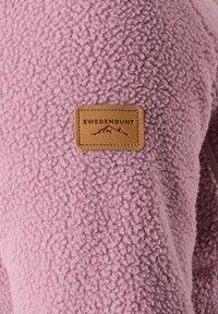 Tissu polaire texturé rose en gros plan avec un patch logo Swedemount rectangulaire en cuir beige représentant un motif montagne.