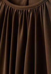 Tela marrón plisada con una textura suave. Presenta puntadas fruncidas a lo largo de la parte superior con pliegues suaves y fluidos.
