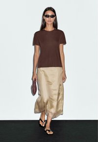 Brown short-sleeve t-shirt, silky beige ankle-length skirt, black flip-flops, carrying a burgundy woven handbag, against a neutral backdrop.