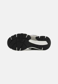 Suela de zapato deportivo negro con goma texturizada, que presenta patrones estriados y un acento blanco cerca de la media suela. Duradera con un diseño de agarre estratégico.