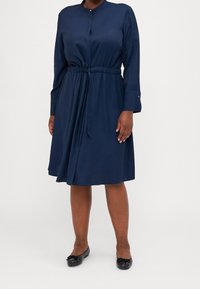 Marineblaues langärmliges Kleid mit kragenförmigem Ausschnitt, tailliertem Bindegürtel und Knopfverschluss. Glattes Gewebe mit einer knielangen Saumlänge.