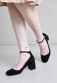 Zapatos de tacón alto de ante negro con punta redondeada, correa al tobillo y tacón cuadrado, que muestran una textura suave y un diseño minimalista.