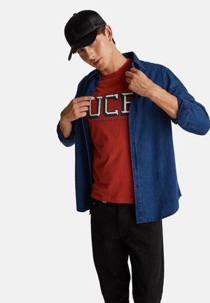 Junger Mann mit schwarzer Kappe, offenem blauem Hemd und rotem T-Shirt mit großen weißen Buchstaben "UCE", der auf das T-Shirt-Logo zeigt.