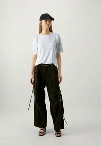 Biała koszulka z kropkowanymi wzorami na rękawach, w połączeniu z ciemnozielonymi spodniami cargo z zamkami błyskawicznymi i sznureczkami, uzupełniona czarną czapką.