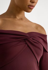 Bordo majica z odprtimi rameni in padajočim dizajnom, z zavihkom na prsnem predelu. Gladek, elastičen material z mat zaključkom.