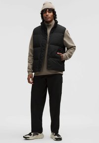 Schwarze Steppweste mit hohem Kragen über einem beigen Reißverschluss-Hoodie, kombiniert mit lockeren schwarzen Hosen und beige/schwarzen Turnschuhen. Weicher Stoff, minimalistisches Design.