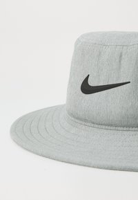 Grå bøttehatt laget av strukturert stoff med en bred brem, med et svart Nike swoosh-logo på forsiden.