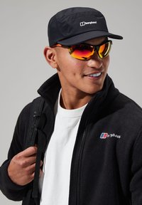 Black fleece jacket, white T-shirt underneath, black cap with logo, colorful reflective sunglasses. Model carrying a backpack.