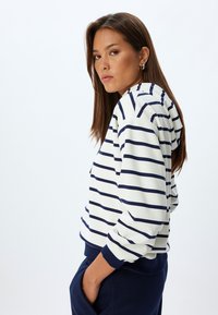 Gestreepte hoodie in crème en marineblauw, met een trekkoordkap, geribde manchetten en een relaxed fit. Het model poseert met de handen in de zakken.
