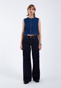 Top corto de mezclilla con dobladillo sin terminar, color azul y diseño sin mangas. Combinado con unos jeans acampanados de tiro alto en azul oscuro que cuentan con bolsillos delanteros.