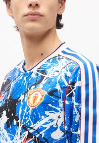 Νέος άνθρωπος που φοράει μπλε φανέλα ποδοσφαίρου της Manchester United με λευκές και κόκκινες ριγέ λεπτομέρειες στους ώμους.