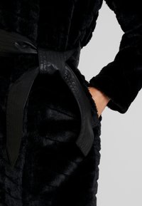 Sort faux fur morgenkåbe med en tekstureret overflade, der har et bredt, fladt sort læderbælte og sidelommer. Maskinvaskbart stof.