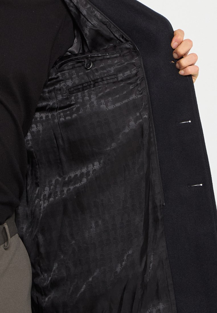 Interno del cappotto nero caratterizzato da un morbido tessuto di satin con un design a motivi discreti, una tasca interna singola e dettagli di cucitura.