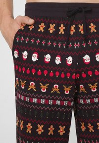 Pantalones de pijama negros con patrones festivos que incluyen figuras de hombrecitos de jengibre, rostros de Santa, bolas rojas y renos, con cintura ajustable con cordón.