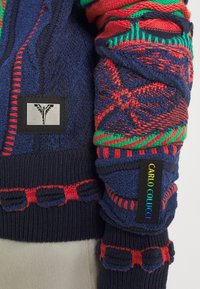 Neulejakku laivastonsinisessä, jossa näyttävät punaiset ja vihreät kuviot. Rakeistetut hihat kontrastivärisellä ompelulla ja logotarrat.