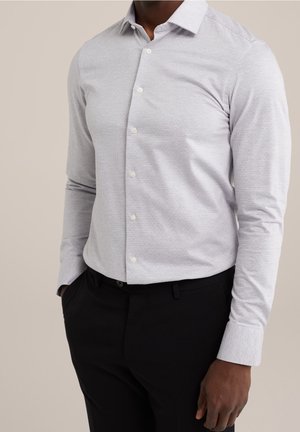 Man wearing a light grey button-up shirt with subtle texture and black trousers, standing with one hand in pocket against neutral background.