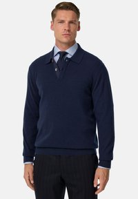 Námořnický modrý pletený svetr s límcem, knoflíkovou légou a žebrovanými manžetami. Nosený přes světle modrou košili s kravatou.