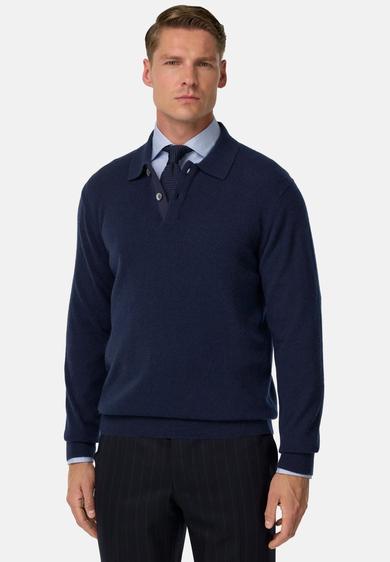 Námořnický modrý pletený svetr s límcem, knoflíkovou légou a žebrovanými manžetami. Nosený přes světle modrou košili s kravatou.