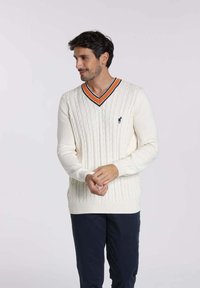 Pull en maille torsadée de couleur crème avec un col en V, rehaussé d'un col rayé noir et orange. Présente un petit logo sur la poitrine.
