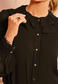 Zwarte blouse voor vrouwen met gerimpelde kraag en hartvormige gouden knopen, met hand die gouden ringen en armband laat zien.