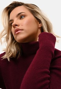 Burgundský rolák s ribovanou texturou a dlouhými rukávy, nošený s zvednutou rukou, ukazující přiléhavý střih. Měkký pletený materiál.