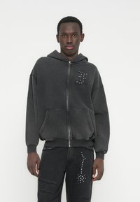 Grauer Kapuzenpullover mit Reißverschluss, Kapuze, großen Fronttaschen und dekorativen Nieten auf der Brust. Kombiniert mit schwarzen, verwaschenen Jeans mit Nietenakzenten.